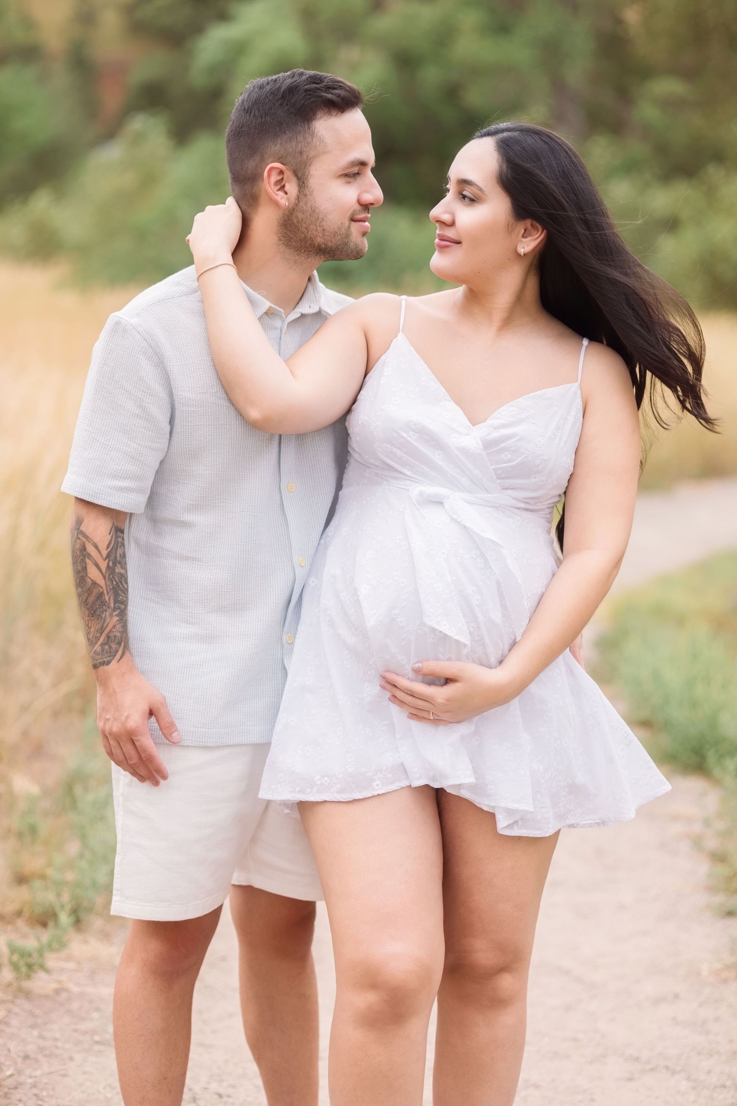 A couple wearing matching white outfits shares intimate moments while walking along a nature path in warm sunlight.