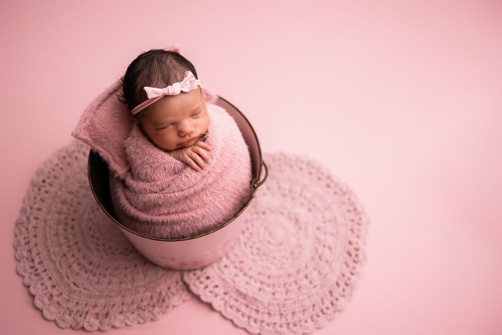 Newborn baby wrapped in pink swaddle sleeping in a wooden bucket on a pink crochet mat with a bow headband.