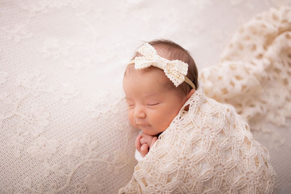 Newborn baby girl swaddled in cream lace wrap with pearl bow headband, sleeping peacefully on soft blanket. Clarkson Michigan photographer