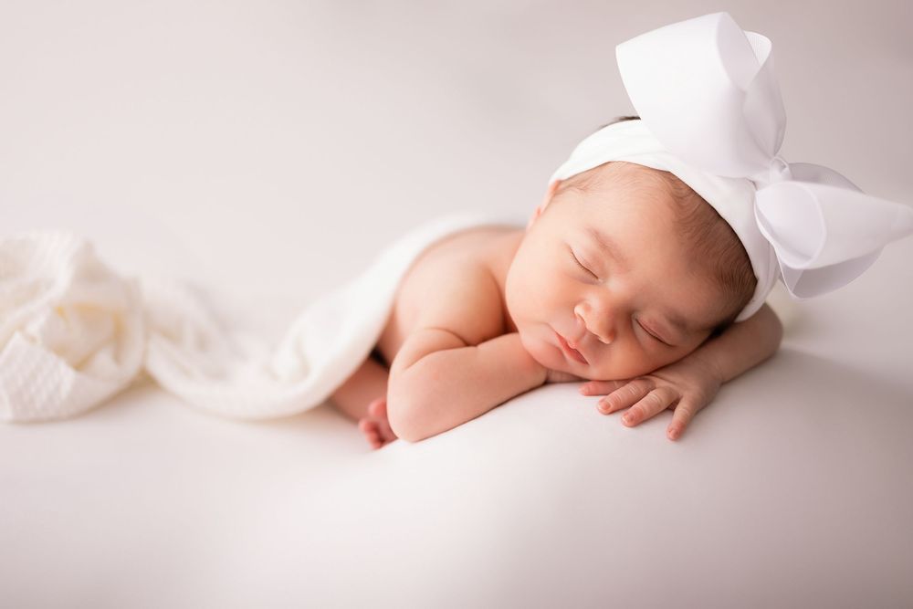 Peaceful newborn baby sleeping with white bow hat and soft blanket in a serene white studio setting. Clarkston Michigan newborn photographer