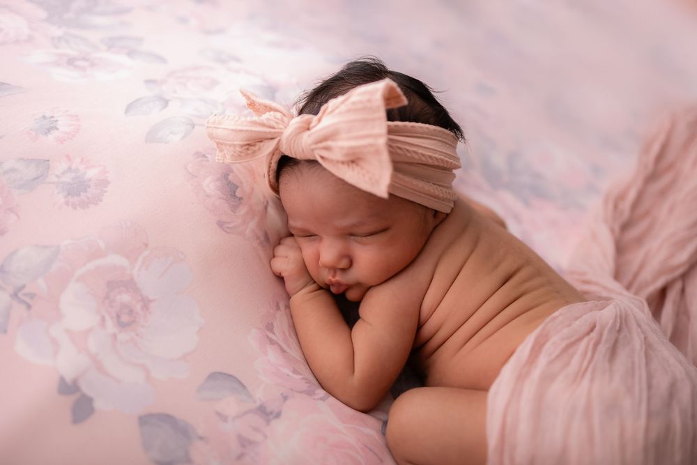 Sleeping newborn baby girl in peach wrap with pink bow headband on floral blanket.