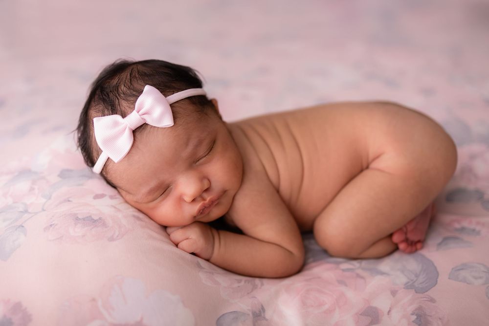 Peaceful newborn baby girl sleeping curled up on a pink floral blanket, wearing a delicate white bow headband.