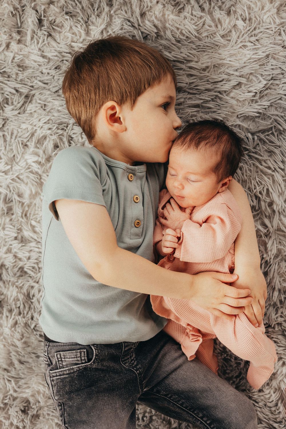 Young boy gently kissing newborn baby sibling while lying together on a soft gray fluffy blanket.