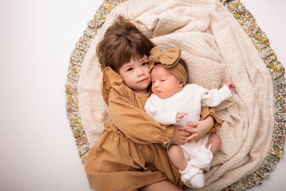 A toddler girl in a brown dress cuddles a sleeping newborn baby wrapped in white on a floral round cushion.