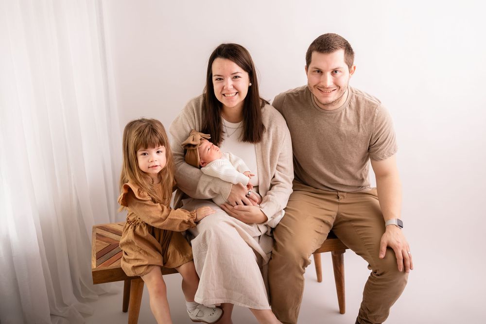 Happy family of four posing for a newborn portrait, parents with toddler girl and baby in bright white studio.