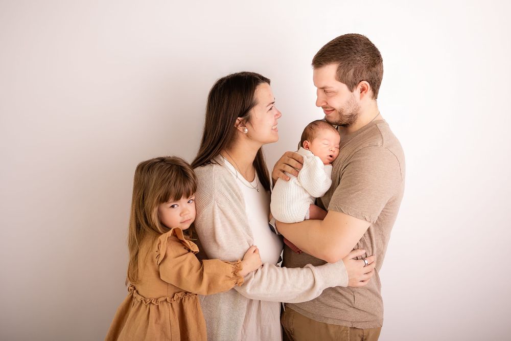 A young family of four poses together, parents lovingly gazing at their newborn baby while a toddler girl stands nearby.