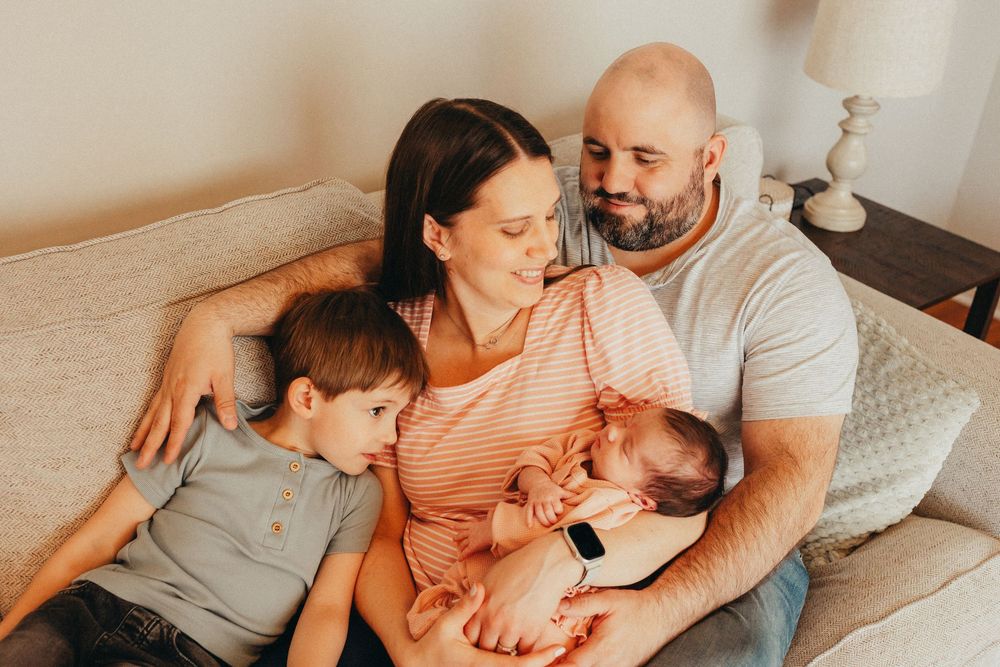 Family of four sitting on couch, parents smiling at newborn baby while young boy cuddles close in warm home setting.