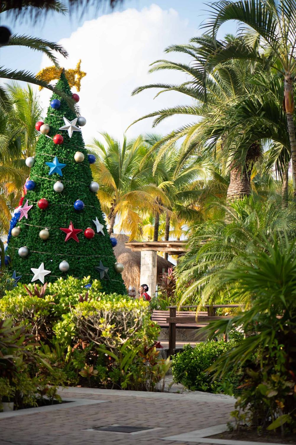 Decorated Christmas tree with colorful ornaments stands among tropical palm trees in an outdoor courtyard setting.