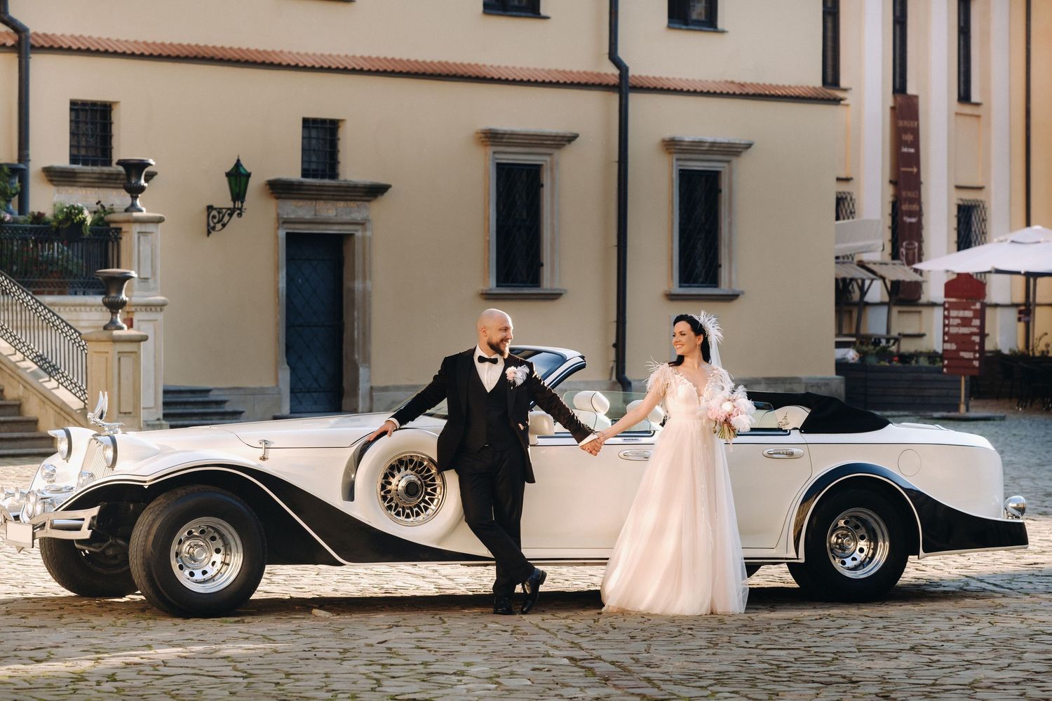 Classic vintage car parked on cobblestone street with formal attire occasion in the background.