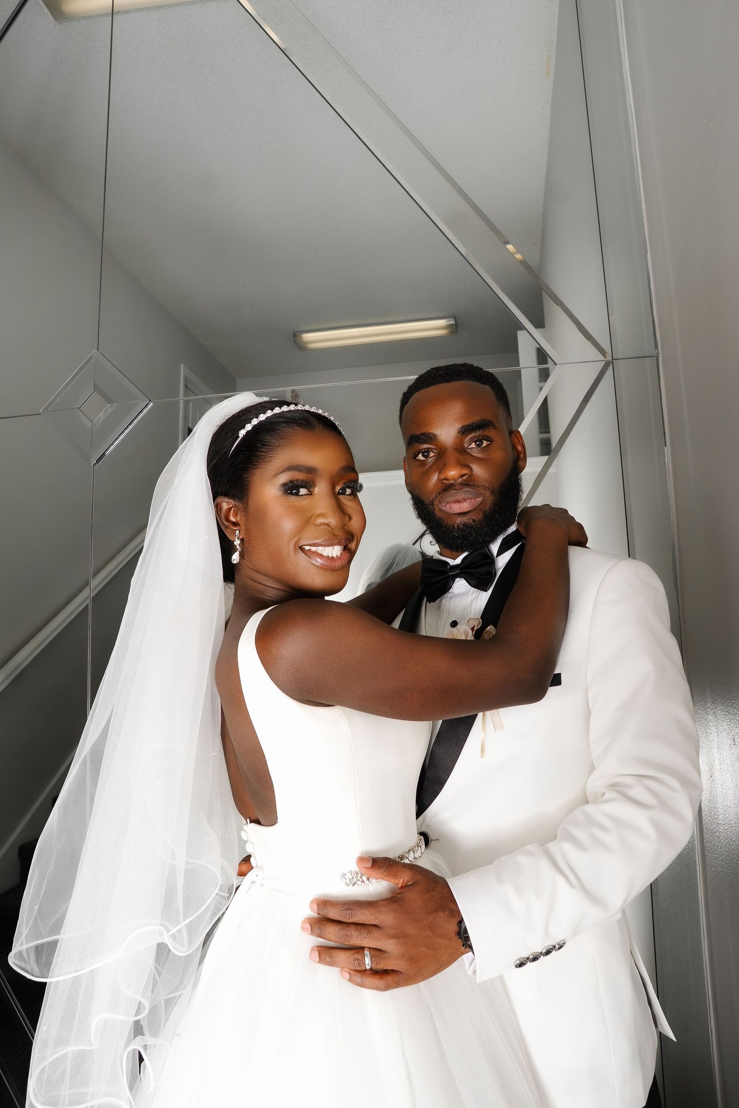 A newlywed couple in elegant wedding attire share intimate moments during a photoshoot in a modern white interior.