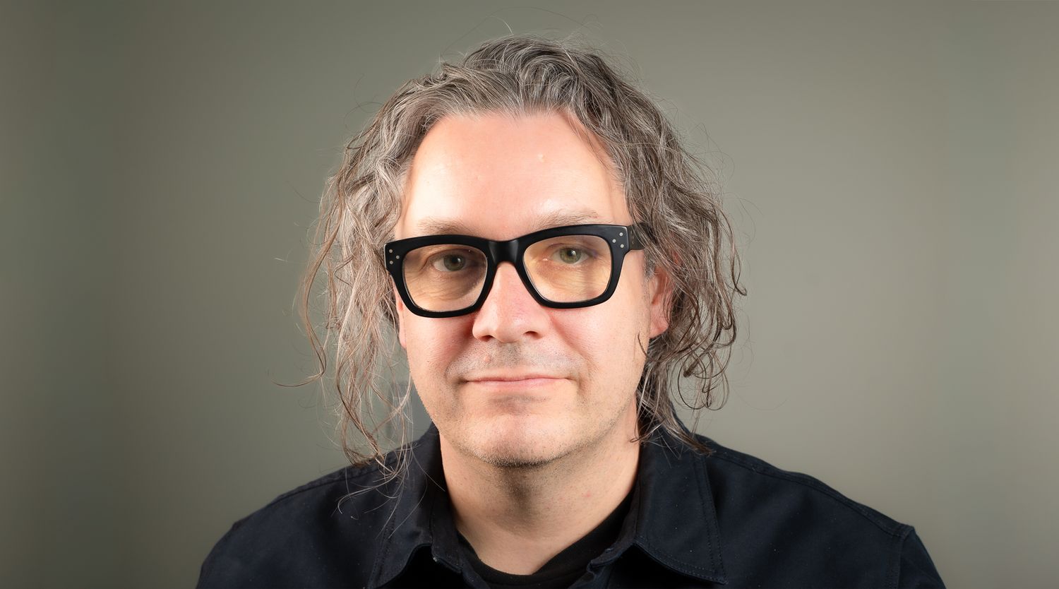 Keith with long brown hair and black thick-rimmed glasses wearing a dark shirt, professional headshot on gray background.