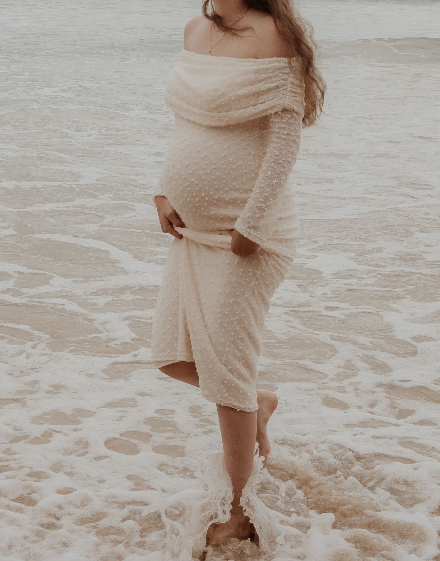 Pregnant lady wearing a cream-colored dress walks barefoot along the shoreline as waves roll in during a central coast maternity photoshoot.