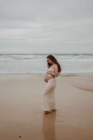 Pregnant woman in a white lace skirt and top stands contemplatively on a misty beach shoreline during a central coast maternity photoshoot.