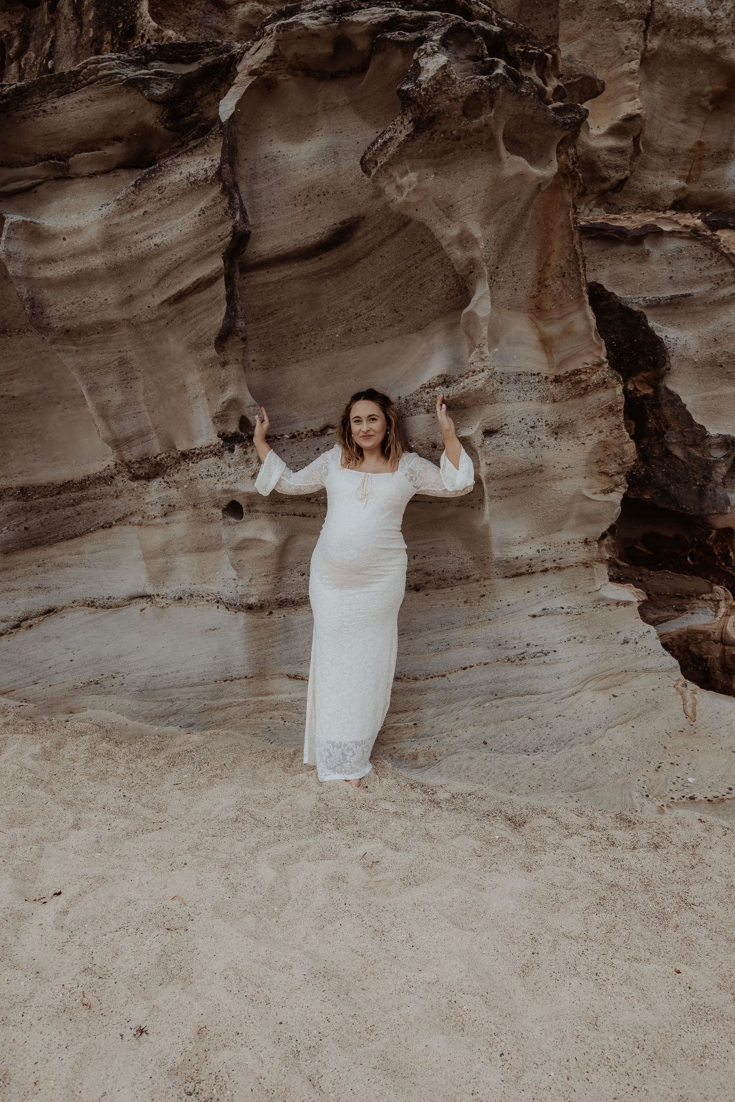 Pregnant woman in white dress standing infront of a rocky coastal cave formation during a central coast maternity photoshoot.