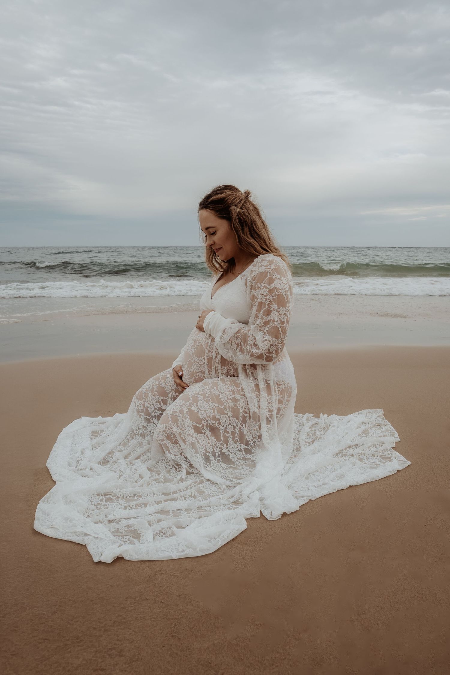 Pregnant woman in white lace dress poses gracefully on beach during overcast day at a central coast maternity session.