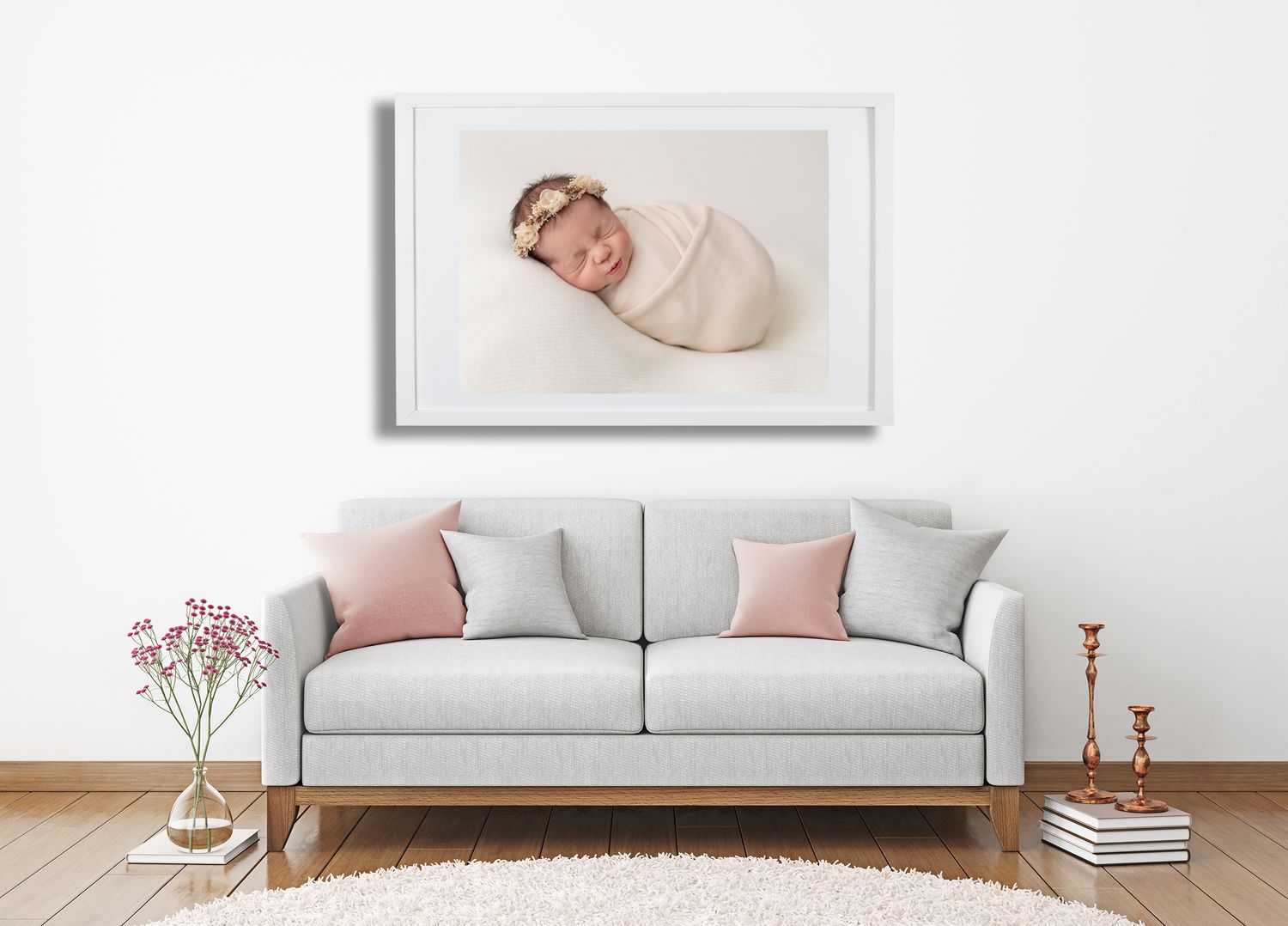 A modern living room interior showcasing a framed newborn photo above a gray sofa, pink accent pillows and white walls.