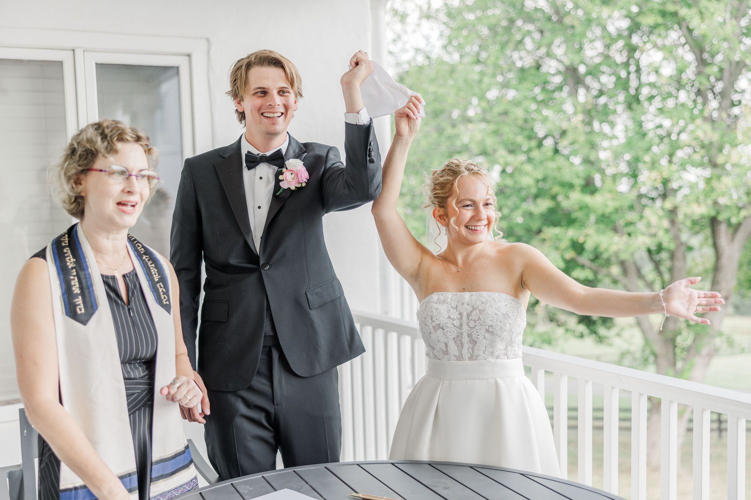 Three people celebrate with raised arms on an outdoor porch during a wedding ceremony.