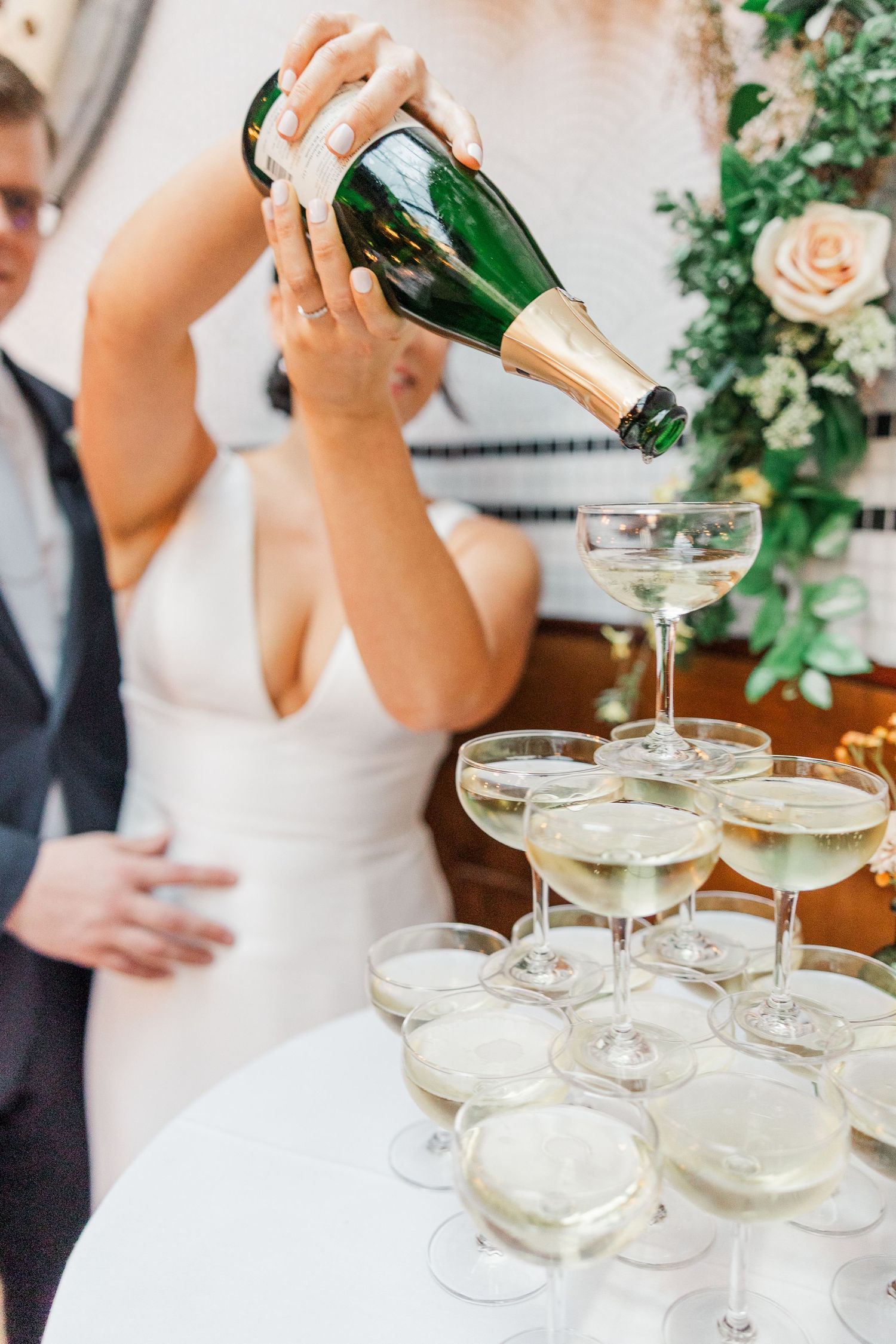 Champagne being poured into a tower of coupe glasses at an elegant wedding celebration.