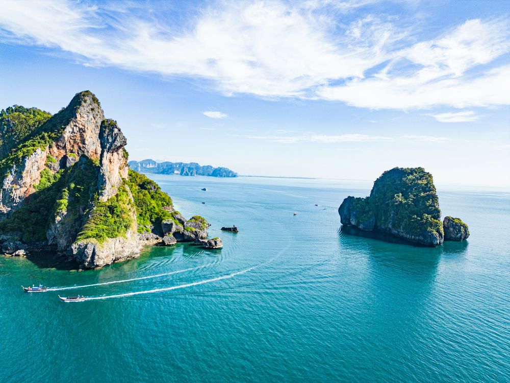 Aerial panorama of limestone cliffs rising from turquoise waters in Krabi, Thailand with long-tail boats cruising below.