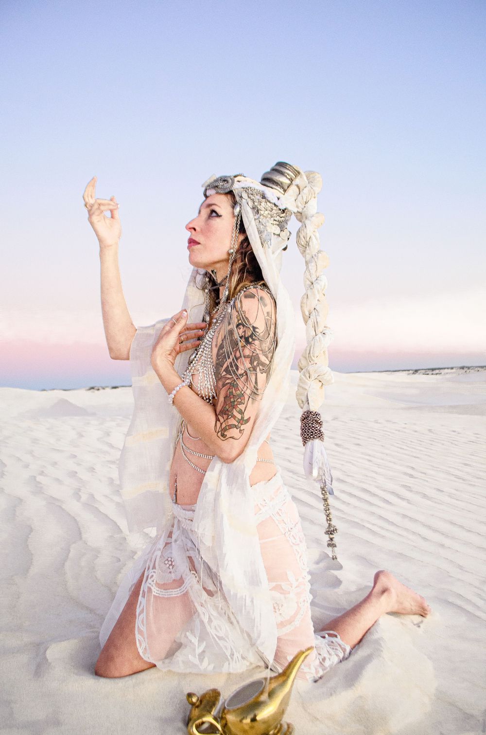 Ethereal figure in white dress poses dramatically on sand dunes at sunset.
