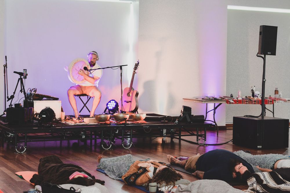 A performer plays acoustic guitar on stage while audience members lie relaxed on the floor during a meditative concert.