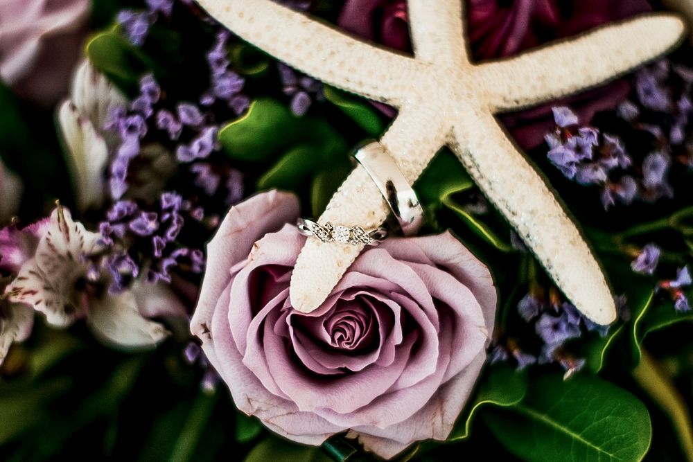 Elegant floral arrangement with dusty purple roses, delicate starfish, and sprigs of purple lavender against dark background.