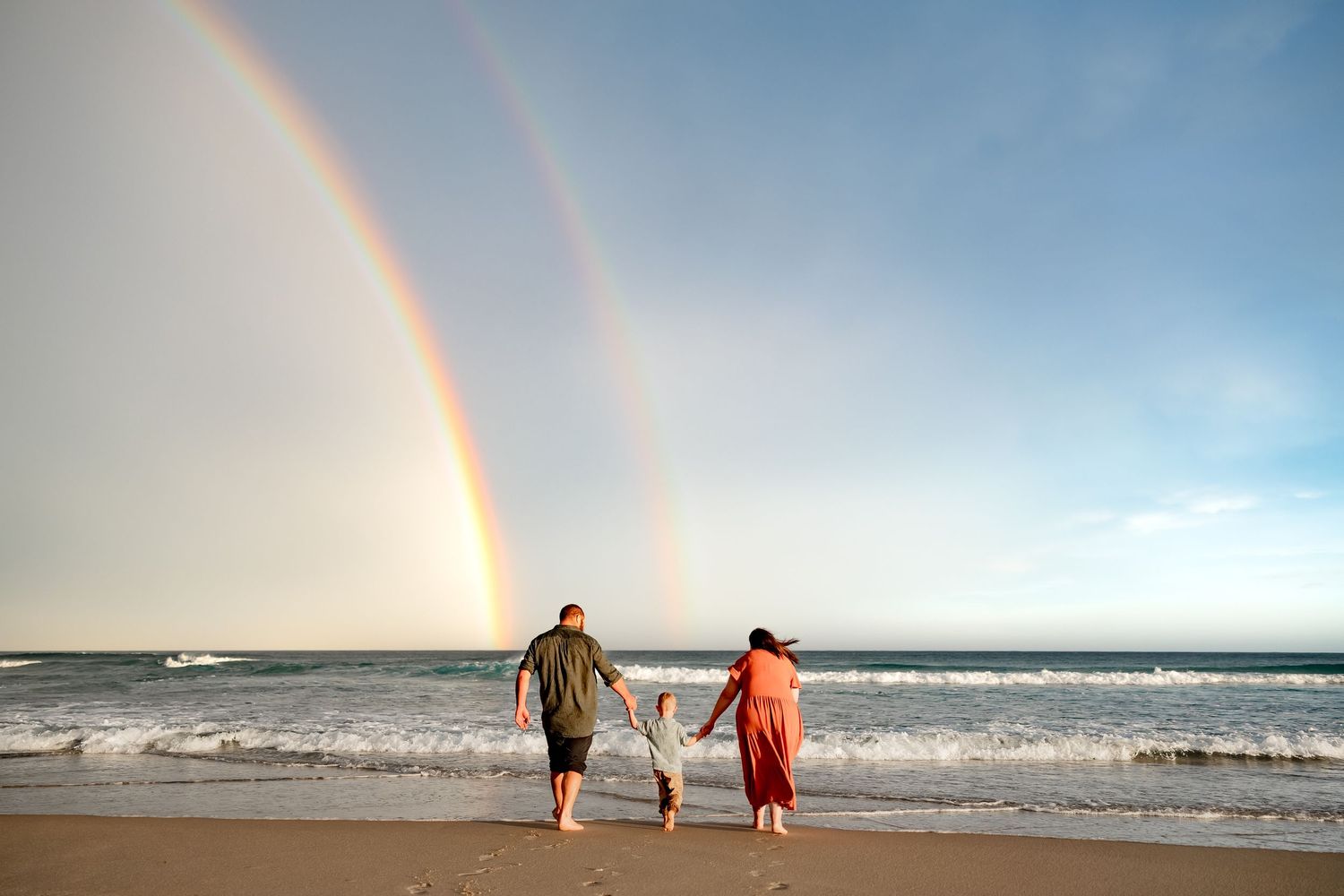 Two people walking hand in hand on a beach while a vibrant double rainbow arches across the ocean sky.