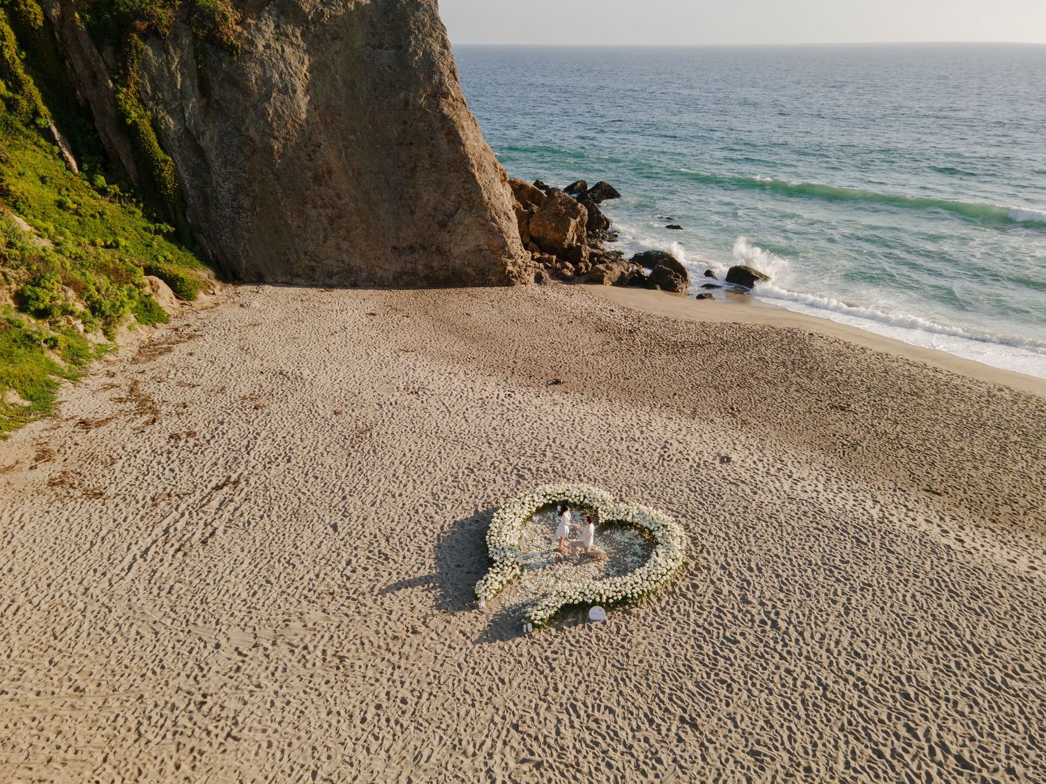 Heart shapes drawn in sand on a rocky beach with turquoise ocean waves and cliffs in the background.