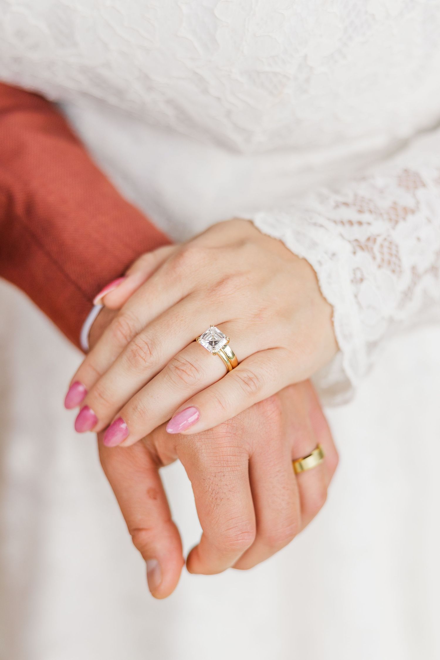 Close-up of hands wearing engagement rings against white fabric showing wedding ring details and pink nail polish.