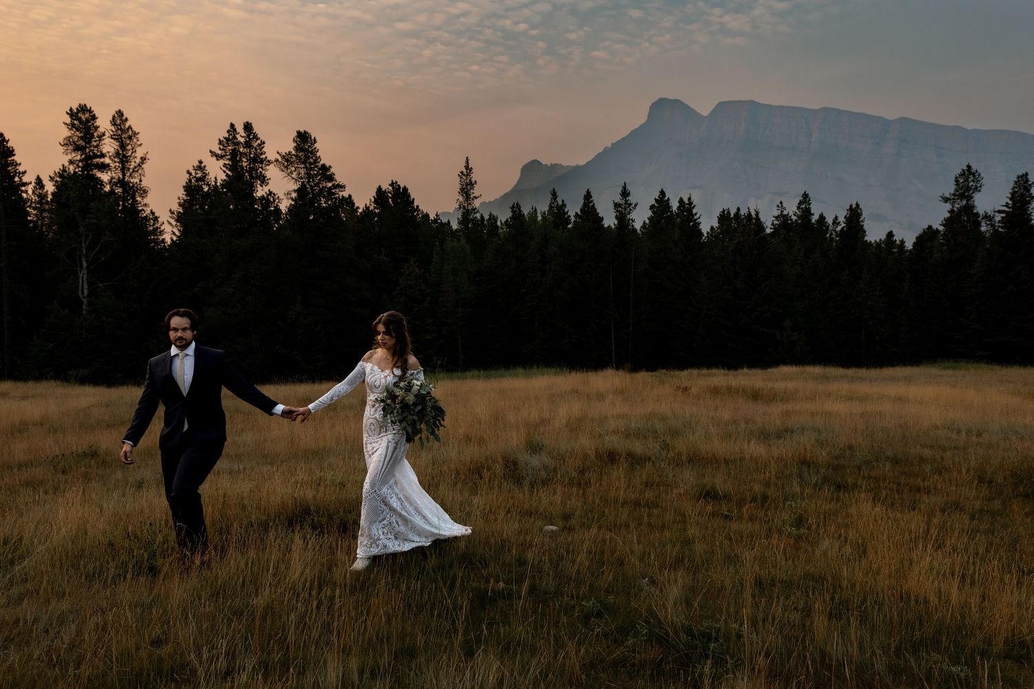A romantic couple in wedding attire walks through a golden field at sunset with mountains in the distance.