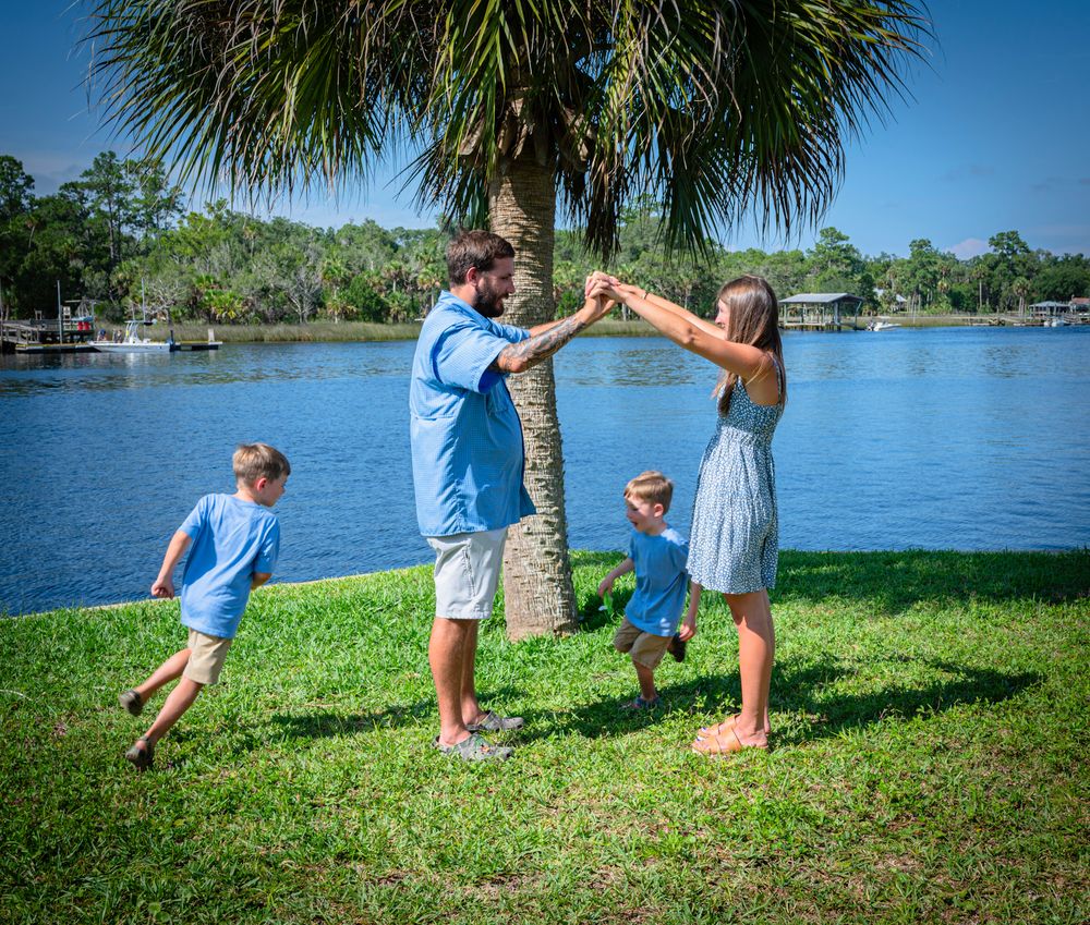 Children play and run near a palm tree by a scenic lakeside on a sunny summer day.