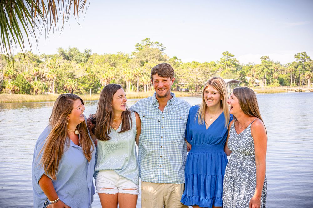 A group stands together laughing and smiling by a scenic waterfront on a sunny afternoon.