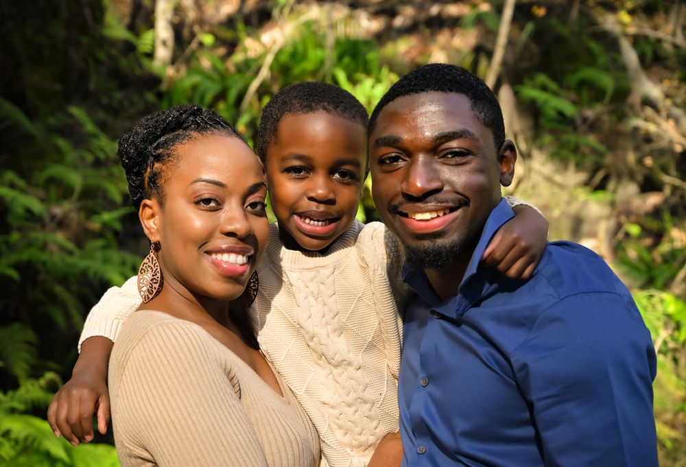 A loving family of three poses together outdoors against a lush green background on a sunny day.
