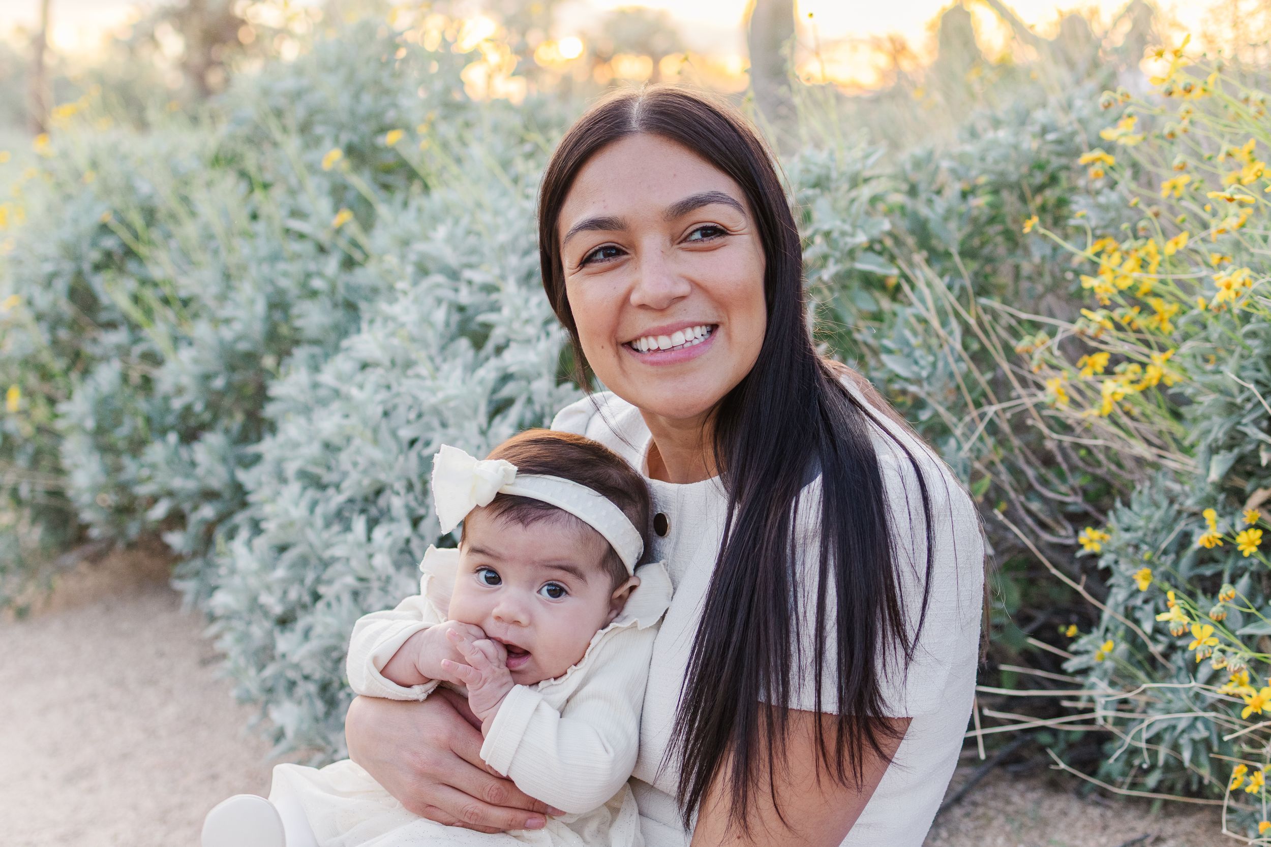 Mom holding infant baby girl next to yellow flowers and light bushes in the middle of the desert during golden hour.