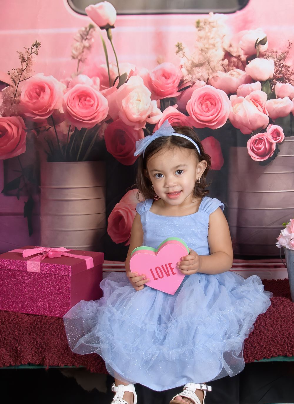 A child in a light blue tulle dress poses with pink roses and Valentine's Day decorations in the background.
