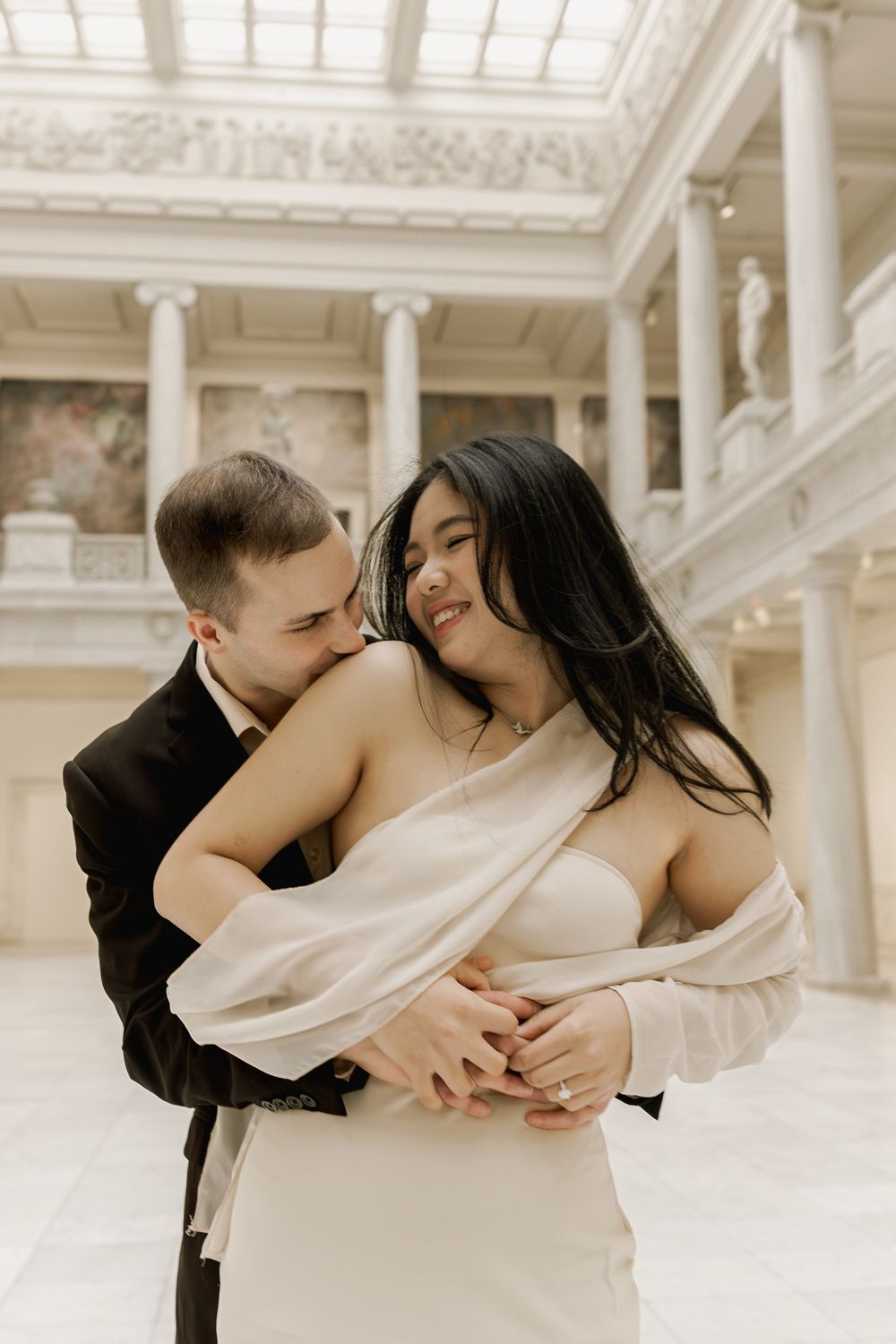 A romantic couple embraces in an elegant white marble hall with columns and ornate architectural details.