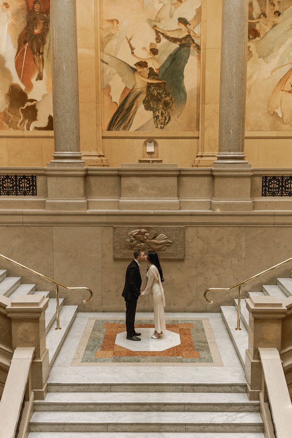 A couple stands on a white marble platform during a wedding ceremony with decorative murals on the walls.