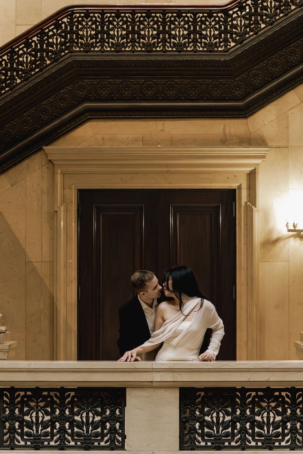 A couple shares an intimate moment on an ornate balcony with decorative railings and architectural details.