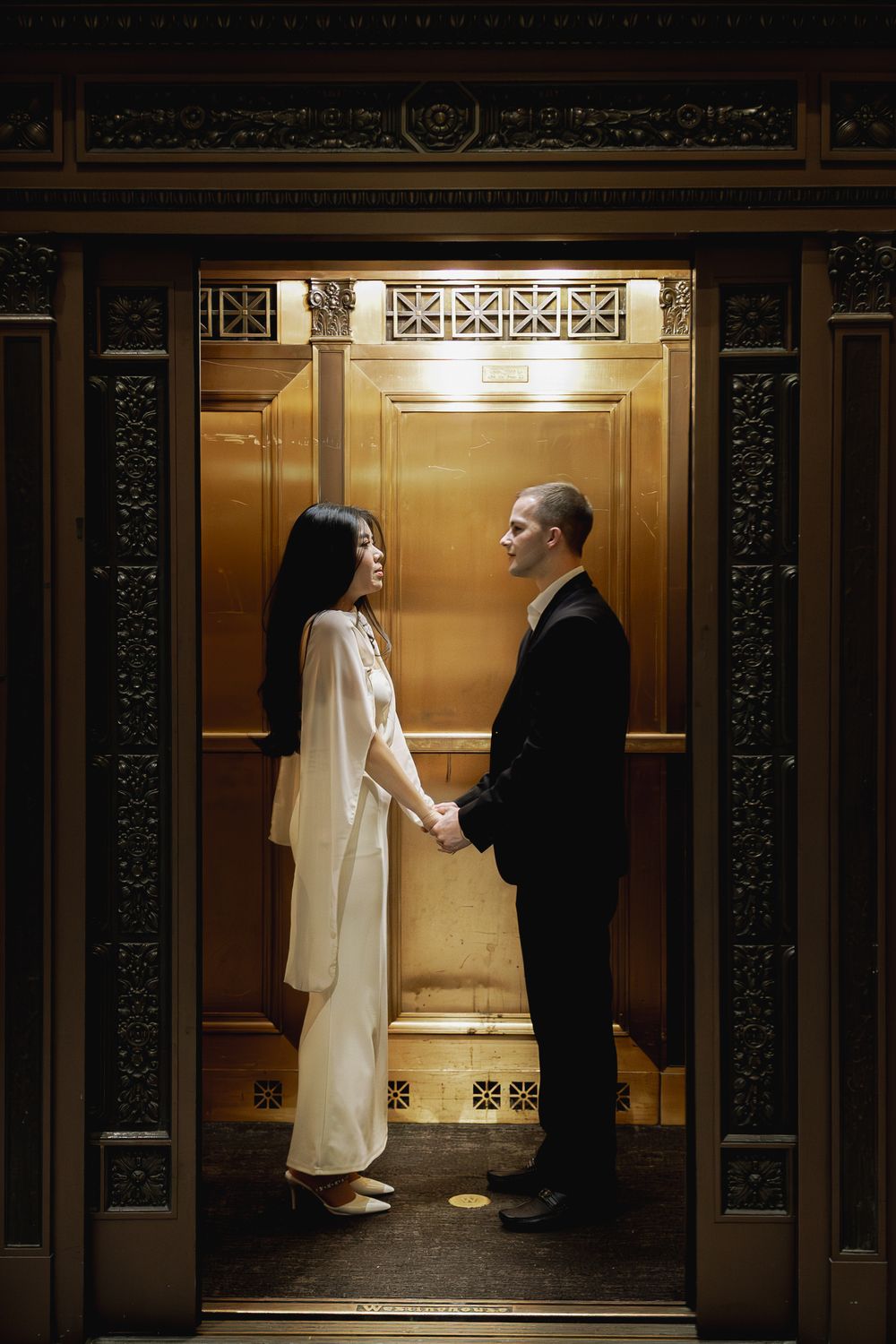 A bride and groom stand together in a vintage elevator with ornate details and warm lighting.