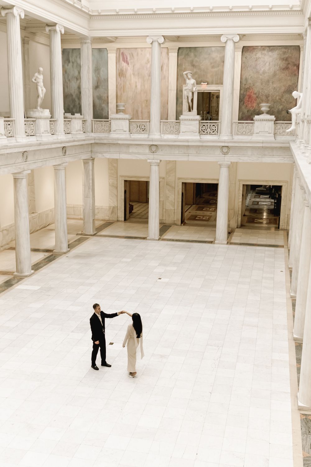 Two people stand in a vast white marble hall with classical columns and balconies in an elegant historic building interior.