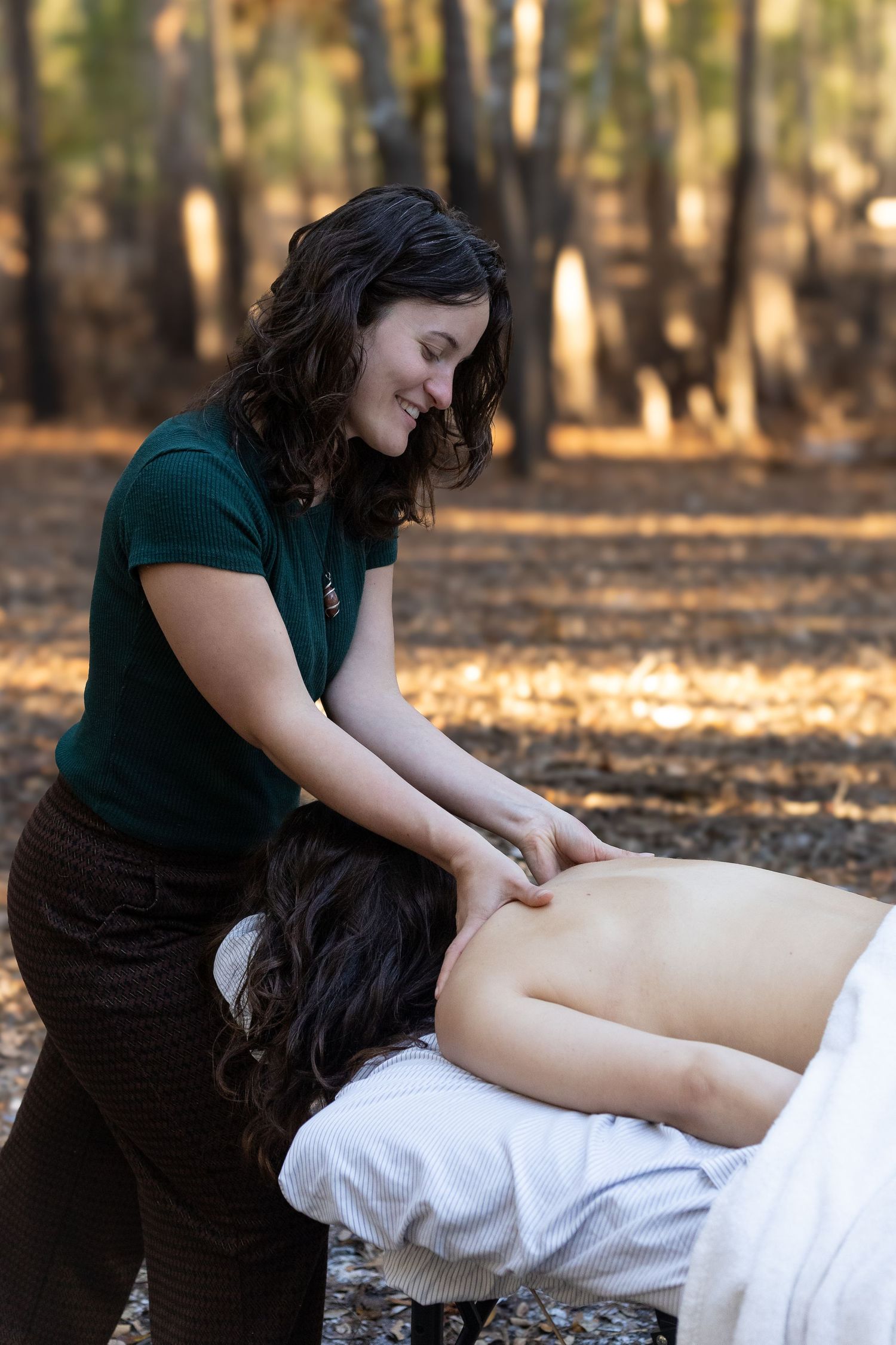 A smiling massage therapist performs an outdoor back massage in an autumn forest setting.