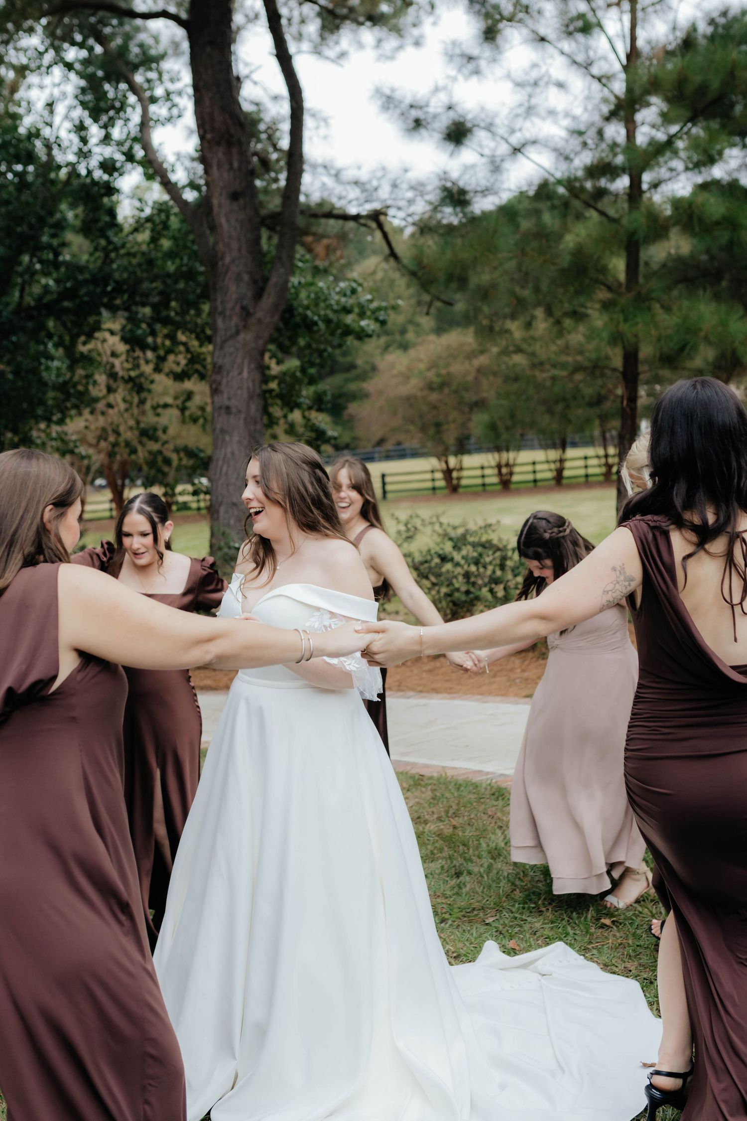 Bridesmaids in brown dresses and bride in white gown hold hands in a line while walking outdoors.