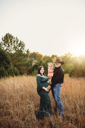 A family maternity portrait in a rustic field during golden hour with autumn foliage.