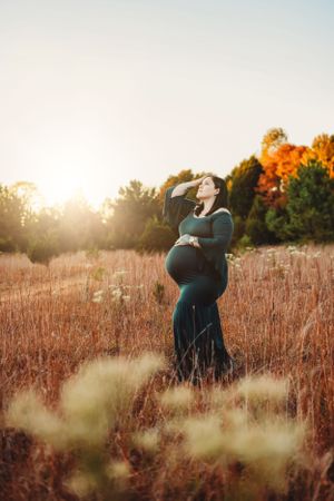 A maternity photo in a golden field at sunset with tall grass and autumn trees in the background.