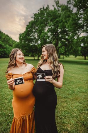 Two women in coordinating mustard and black dresses share ultrasound photos outdoors.