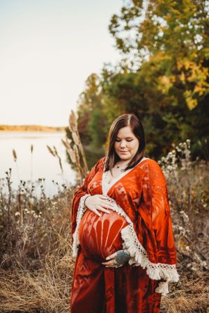 Maternity photo by a lake featuring a flowing red dress with fringe details at sunset.