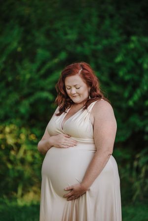 A maternity photoshoot in a lush green outdoor setting features a cream-colored dress.