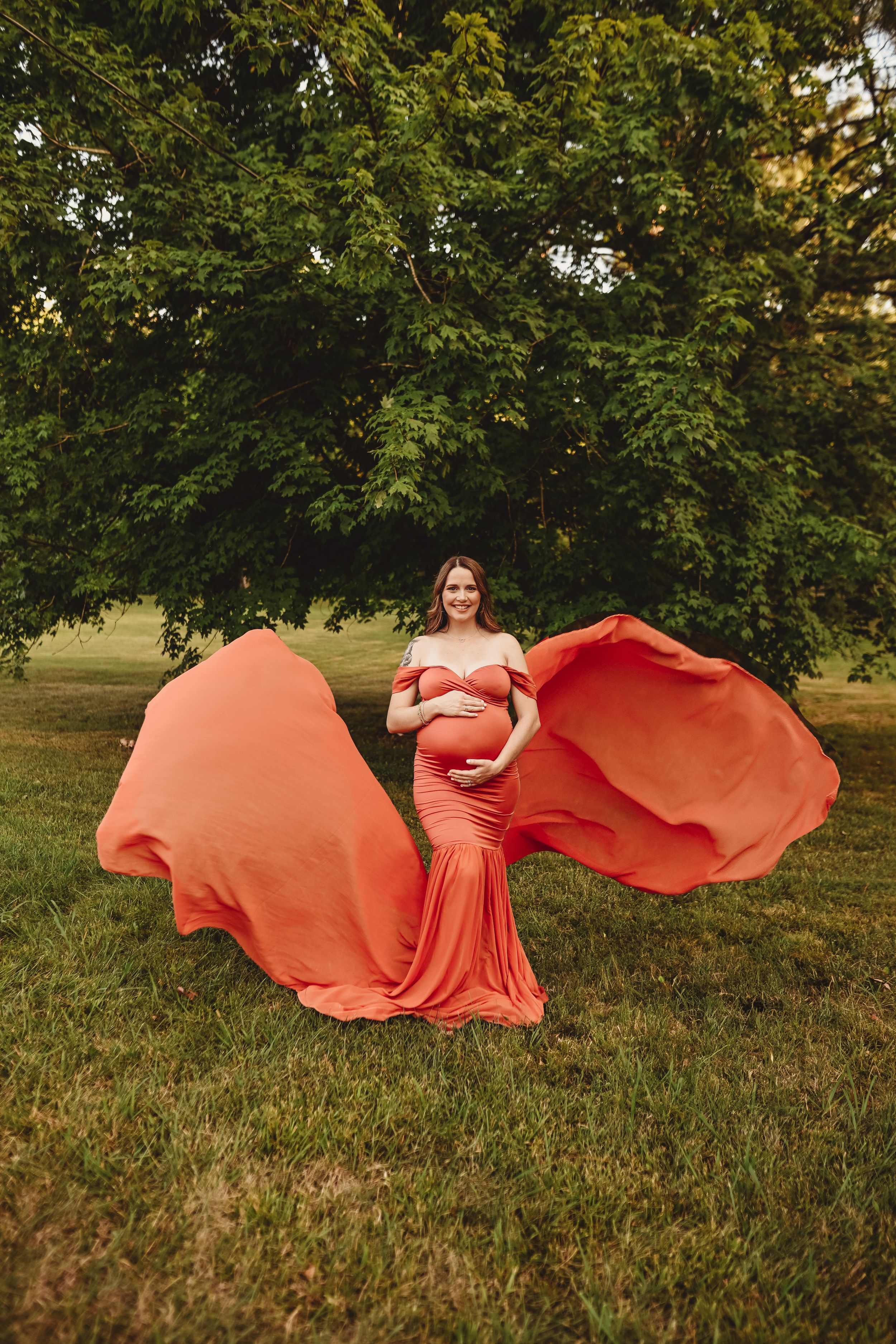 A dramatic maternity portrait with a flowing coral dress against a green landscape.
