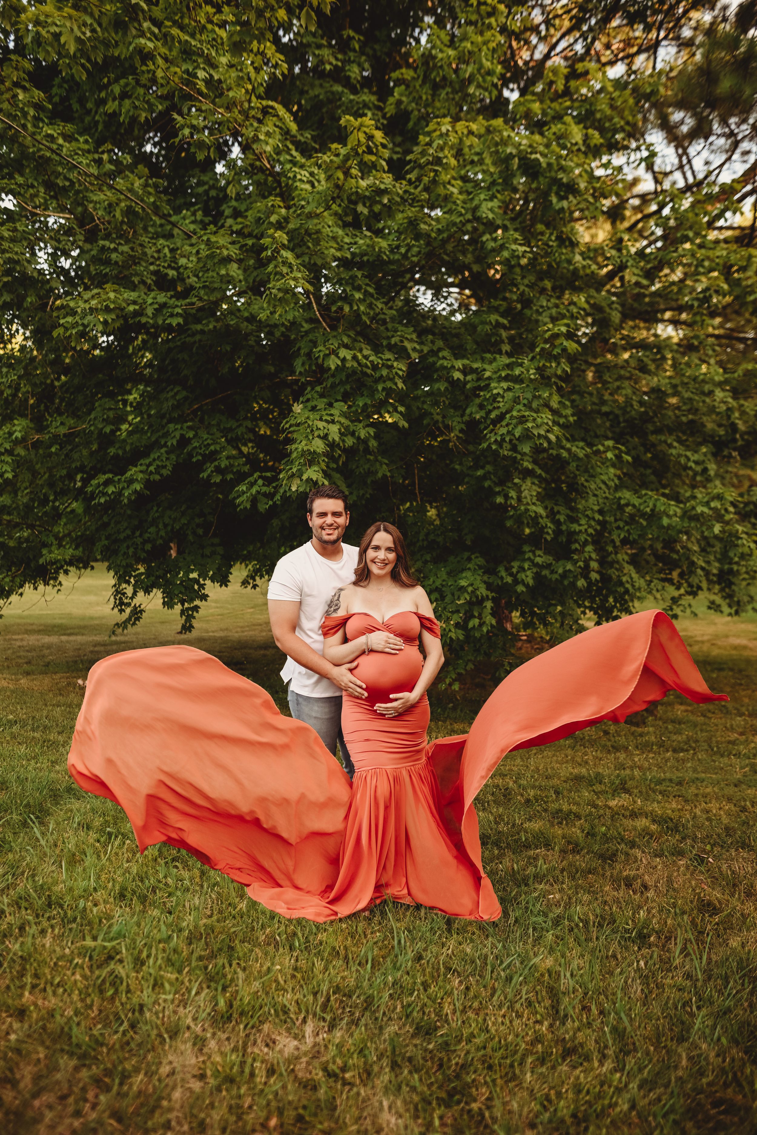 A maternity photo featuring a flowing coral dress billowing in the wind under a large tree.
