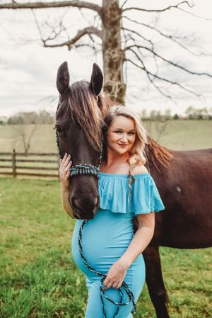 A close-up shot of a dark horse with someone in turquoise dress standing in a fenced pasture.